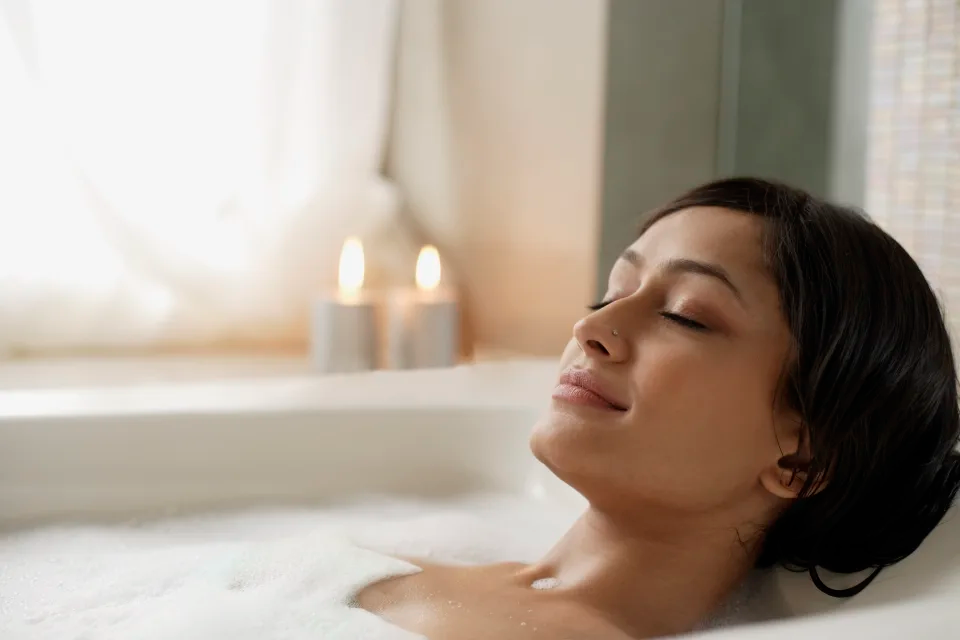 Woman with eyes closed, relaxing in a bubble bath by a window. 2 candles are lit on the window sill.  
